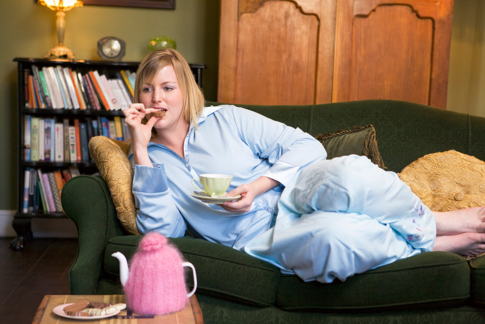 A young woman lying on her couch drinking tea and eating cookies to illustrate How To Stop Snacking After Dinner