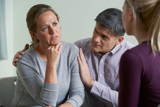 A man comforting a woman who looks worried as she talks to a therapist (back to the camera)