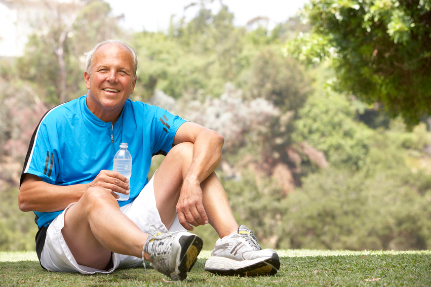 A smiling older man with gray hair, wearing a t-shirt, shorts, and running shoes sitting down in a park with a bottle of water to illustrate sustainable weight loss.